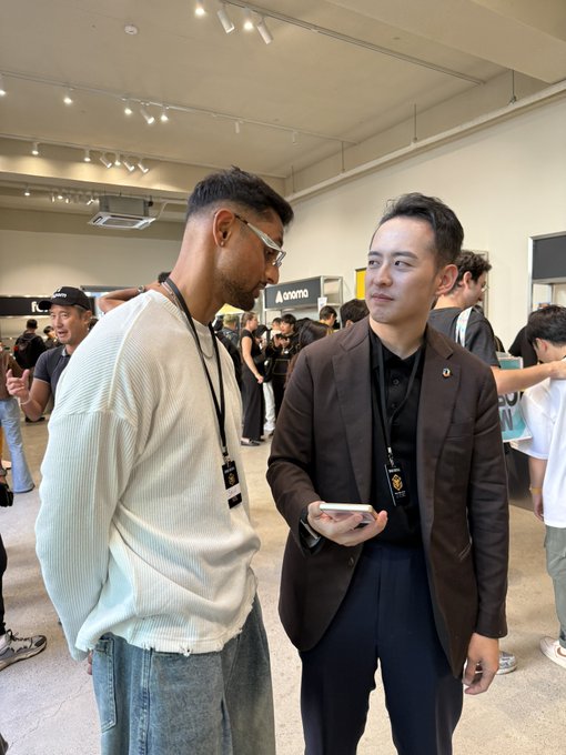 Two men conversing at what appears to be a crypto or tech conference, one wearing sunglasses and a white sweater, the other in a black blazer holding a phone, both wearing event lanyards.