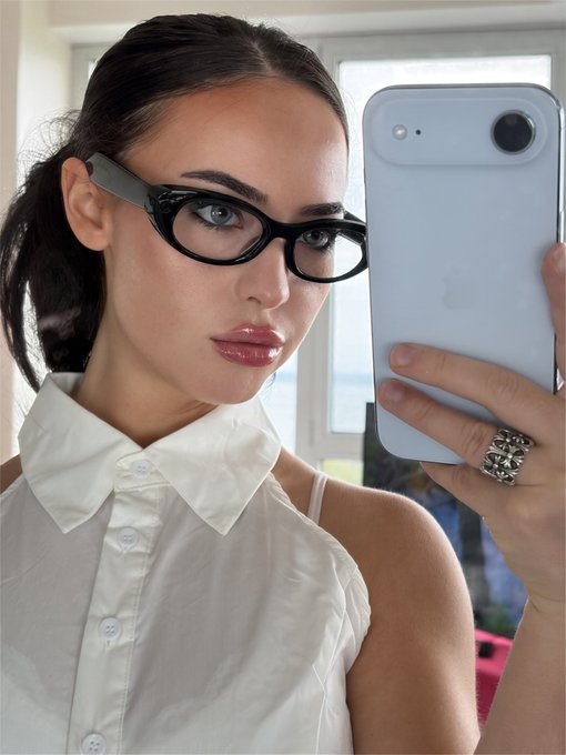 A young woman with dark hair pulled back takes a mirror selfie wearing black-framed glasses and a white sleeveless button-up top, holding a light blue iPhone adorned with a chunky silver ring. The polished, confident aesthetic complements the post's "Good morning super intelligent hard money investors" greeting, projecting a put-together professional vibe fitting for a financial audience.