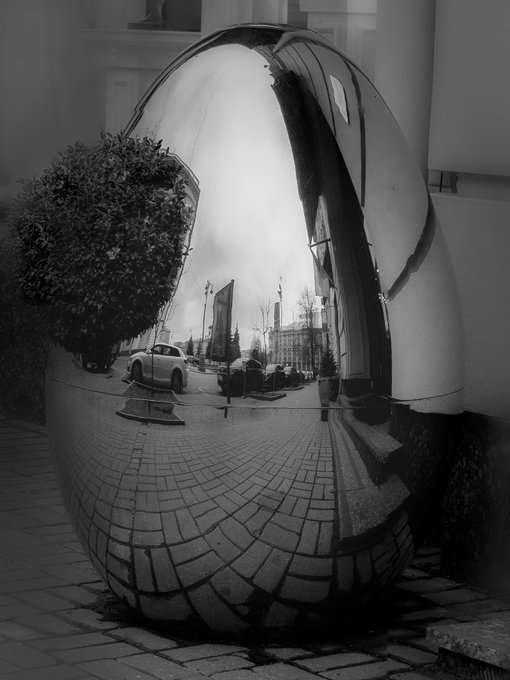 A reflective metallic egg-shaped sculpture sits on a cobblestone surface, its mirrored surface capturing a distorted, fish-eye view of a street scene with a white car, buildings, and an overcast sky. The image relates to the listed artwork series, likely showcasing one of the limited-edition sculptural pieces, where "Geometry of Transience" appears to correspond to this mirrored form that warps reality through its reflective geometry.