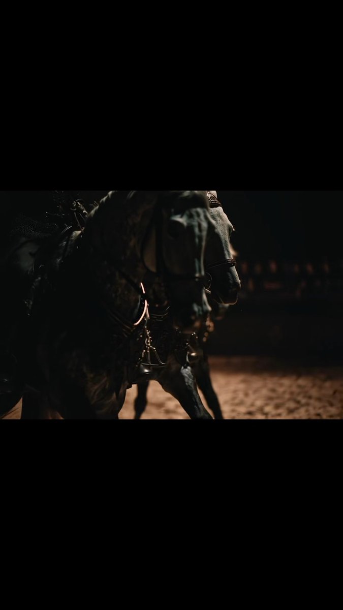 A dark, cinematic close-up of a horse in motion, with light catching its bridle and bell against a sandy arena background.