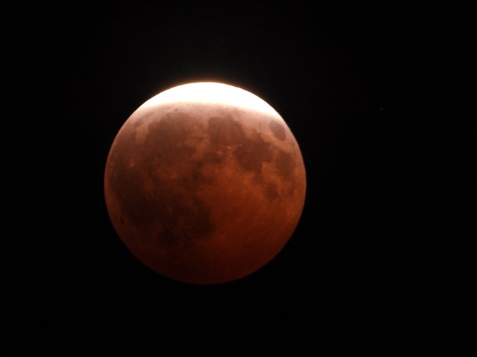 A striking photograph captures a lunar eclipse, with the moon displaying its characteristic deep reddish-orange hue across most of its surface while a bright white sliver of sunlight illuminates the top edge against the dark night sky.