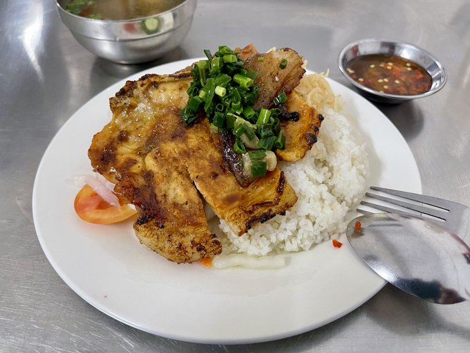 A plate of food featuring grilled chicken with green onions, served over white rice with a side of dipping sauce and a small bowl of broth, accompanied by a fork and spoon on a metal table.