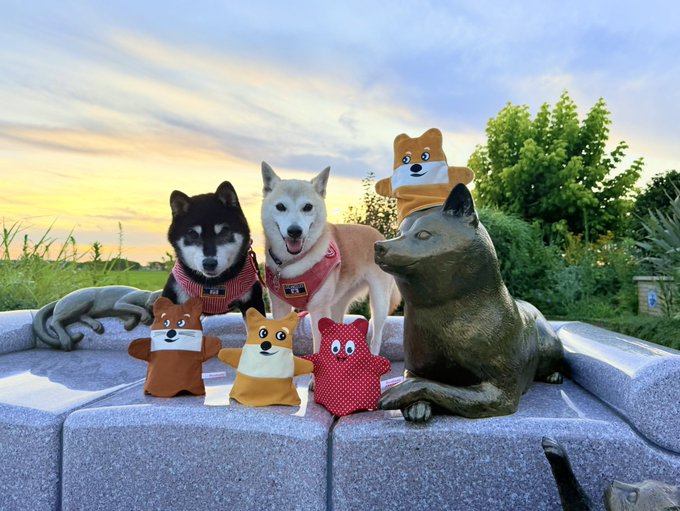 The image features a group of five dogs, including two black ones and three brown ones, sitting together with their heads tilted upwards as if posing for a photo. The dogs are accompanied by four stuffed animals, one red and one yellow, which appear to be part of an art project or a display at an auction. The background includes trees and bushes, creating a natural setting for the scene.