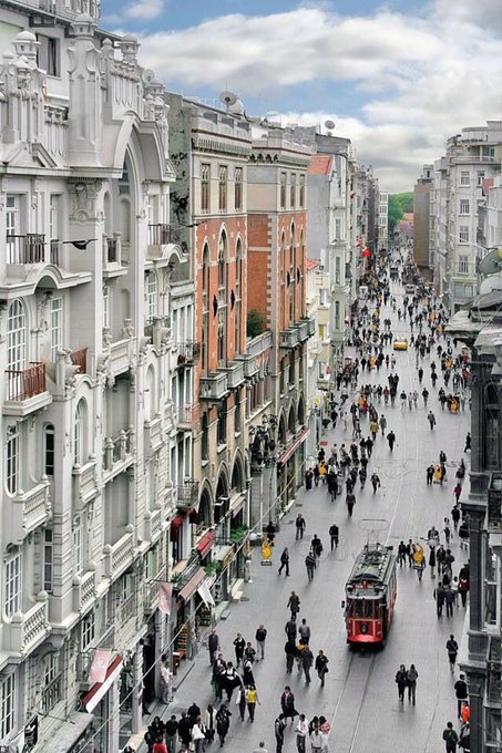 The image shows a busy city street with people walking along the sidewalks, some carrying handbags or backpacks. A red trolley car can be seen moving down the street, adding to the bustling atmosphere of the scene. In the background, there are tall buildings lining both sides of the street, giving it an urban feel. The image also contains a caption that reads "GM CT Not enough people are talking about how @NomismaNetwork ties access to verifiable history Your wallet score determines your mint cost determines your Diamond earnings That'S the foundation everything is built on."