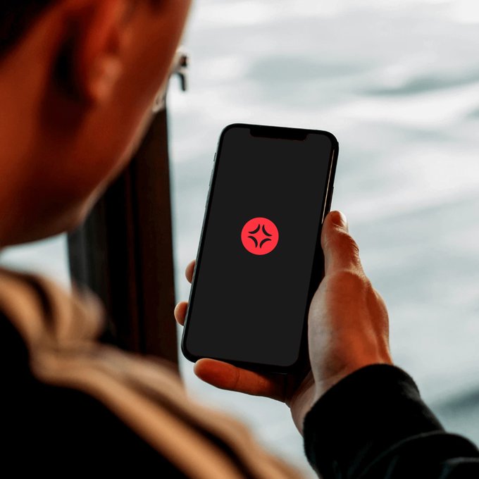 A person gazes at their phone displaying a red circular logo with a four-point star, set against a calm water backdrop, as they check the markets in line with the post about early-morning chart watching.