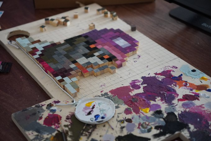 An artistic workspace showing a pixelated portrait painting on a grid board alongside a paint-covered palette, with wooden cubes arranged to form the image.