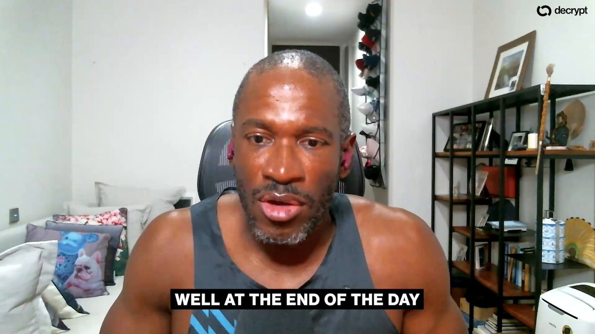 The image shows a man wearing a black tank top sitting at a desk with a computer monitor in front of him, appearing to be talking or giving a presentation. The background features a white wall with a shelf containing various items such as books and bottles. There is also a chair visible near the desk.