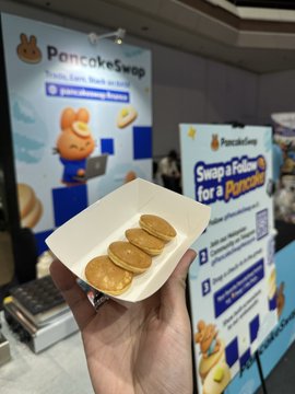A hand holding a paper tray with three small pancakes, with a 'PancakeSwap' banner reading 'Swap a Token for a Pancake' in the background.