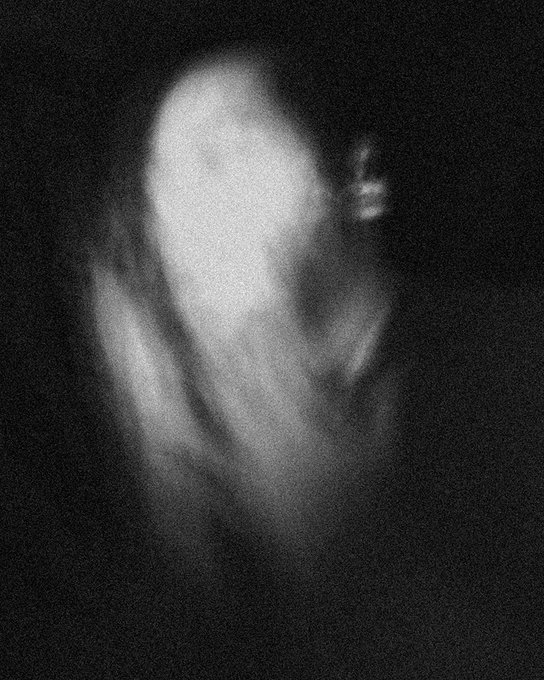 A blurry, grainy monochrome figure appears as a ghostly silhouette dissolving into a black void, evoking a sense of fading presence. This haunting visual aligns with the title "self portrait fading into silence," capturing the intangible and ephemeral quality of identity dissolving into absence.