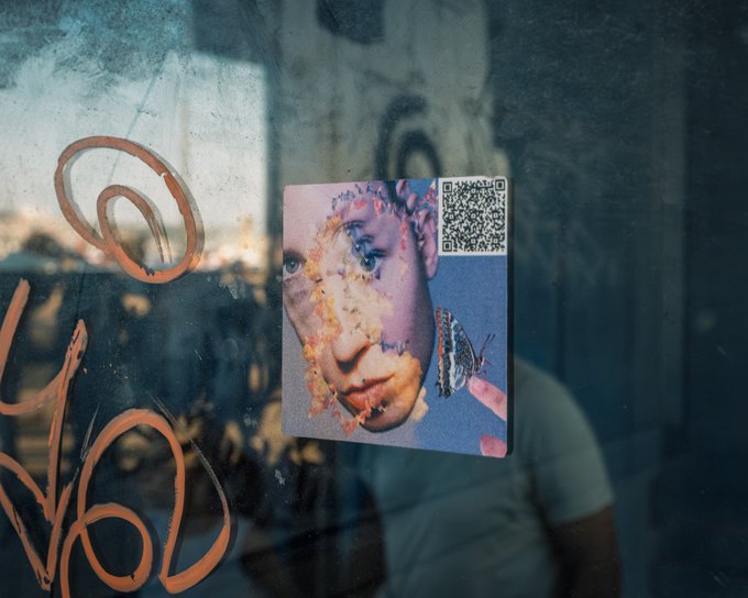 An artistic photograph of a small portrait or photo of a person's face with a QR code, attached to a reflective, textured surface with orange graffiti-like markings.