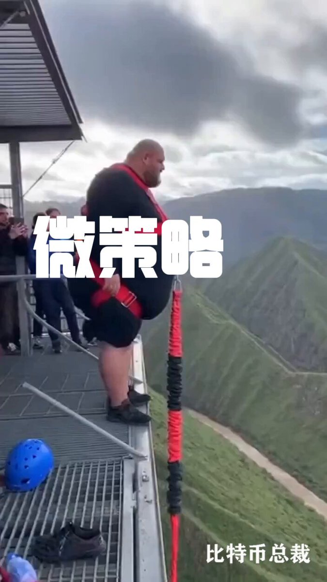 The image shows a man standing at the edge of a cliff with his legs dangling over the side, wearing a harness that has Chinese characters on it. The scene appears to be set in an outdoor location, possibly near a mountain or hillside. In the background, there are several people who seem to be watching the man's daring feat from below. The post includes text written in Chinese and English, which reads: "How devastating is MicroStrategy' s coin-selling? It'S been almost a week, and the crypto circle still haven't recovered."