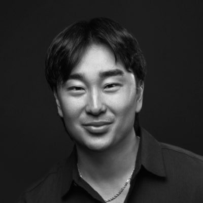 A black and white portrait of a smiling man wearing a dark shirt and a necklace.