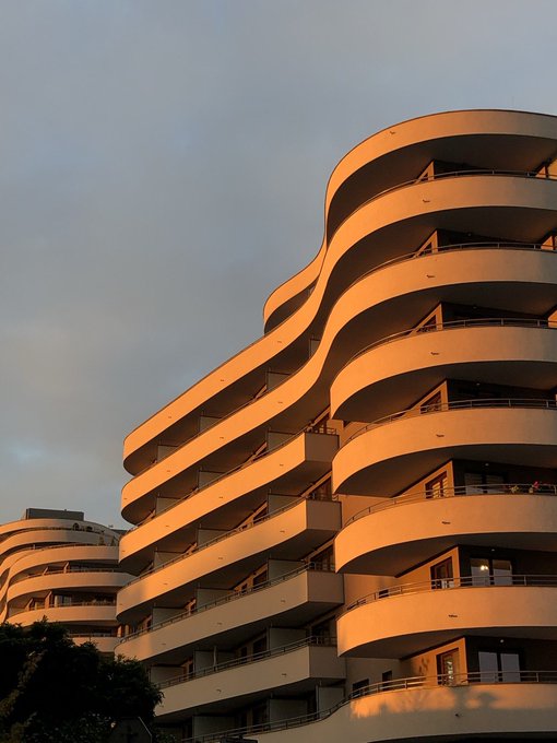 A photograph of a large building with curved, stacked balconies illuminated by warm orange sunset light against a cloudy sky.