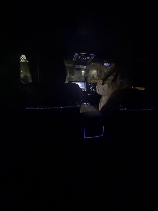 Interior view of a car at night showing a person in the driver's seat with city lights visible through the windows.