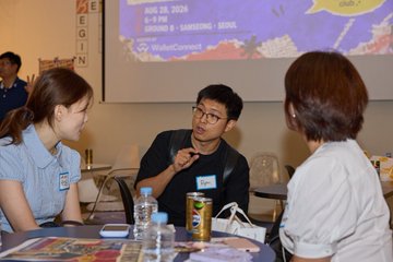 Three people sit at a table during an event with a presentation screen in the background displaying 'AUG 28, 2026, 6-9 PM, HOMME B • SAMSUNG C VENUE, WannaCupBKK'.