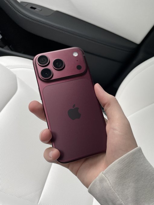 A close-up photo of a hand holding a burgundy/dark red iPhone, likely an iPhone 17 Pro or similar model with a distinctive camera island, against a white car interior background.
