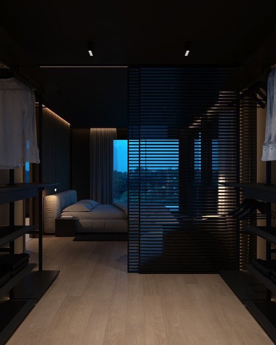 The image shows an open walk-in closet with a bed inside of it. The room has a modern design featuring black blinds that allow natural light to enter, creating a serene atmosphere. A window can be seen through the blinds, providing a view of trees outside. The photo is taken from within the closet, giving a sense of being in the space. The image also includes text about GM @useTria virtual card and how it works, which seems unrelated to the main subject matter of the closet.