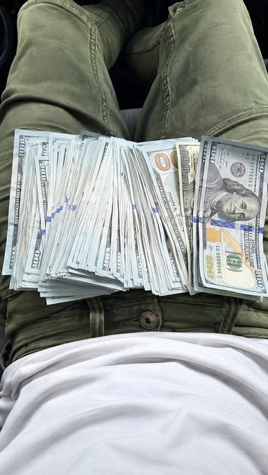 A person rests a fanned-out spread of $100 bills across their lap, with the serial numbers "HB 84658671 J" visible on the top bill. The image visually represents the post's theme of "Manifesting this," serving as a tangible expression of the user's aspiration to accumulate wealth.