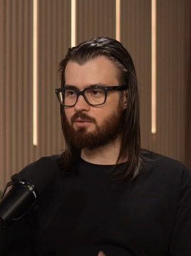 A man with long dark hair, a beard, and glasses speaks into a microphone during what appears to be a podcast or interview, likely discussing the significant $52M loss in his memecoin portfolio mentioned in the caption.