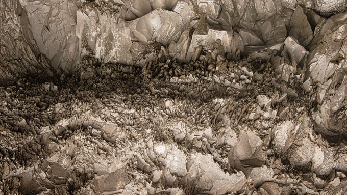 A textured 3D rendering of a rocky, uneven surface with layered stone formations in earthy brown and beige tones, possibly a canyon wall or quarry interior.