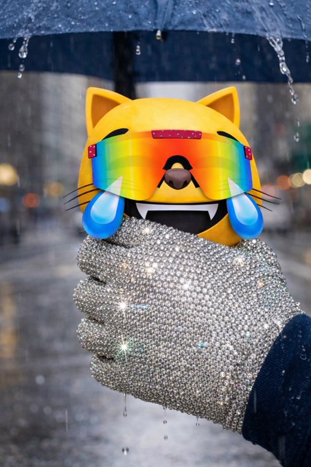 A whimsical image featuring a yellow cat emoji face with rainbow sunglasses and crying laughing tears, held by a hand covered in rhinestones under a rainy umbrella.