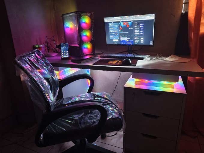 A dimly lit gaming setup features a RGB-illuminated PC tower with three fans, a wide monitor displaying graphs and data, and a holographic chair, with a phone showing "12:46" propped on the desk. The post "My setup Almost Complete - How is it?" invites feedback on this near-finished battlestation.