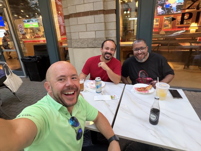 The image shows three men enjoying pizza and drinks at an outdoor table, which aligns with the post about a good time catching up. Key text visible on the bottle in the foreground includes "Root Beer".