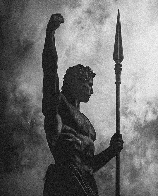 A dramatic black-and-white silhouette of a muscular warrior or statue holding a spear, with one arm raised in a fist against a cloudy, atmospheric background.