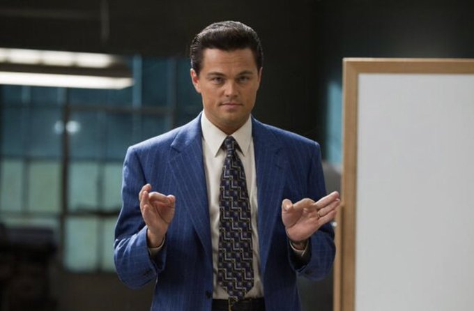 Leonardo DiCaprio as Jordan Belfort in a scene from The Wolf of Wall Street, wearing a blue suit and patterned tie while gesturing with his hands in an office setting.
