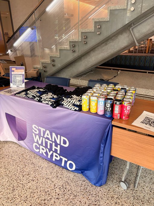 A table display with a purple 'STAND WITH CRYPTO' banner, merchandise t-shirts, and various colorful beverage cans next to a staircase.