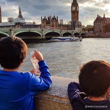 Two boys lean against a railing by the River Thames, looking toward Big Ben and the Houses of Parliament in London, with a watermark "@watchmewsx".