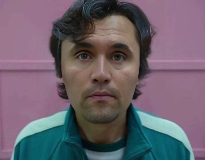 A close-up shot captures a man with dark wavy hair and a solemn expression, wearing a teal tracksuit with white stripes against a pink wall — evoking the likeness of a character from a popular dystopian drama, mirroring the tense anticipation of a high-stakes wager.
