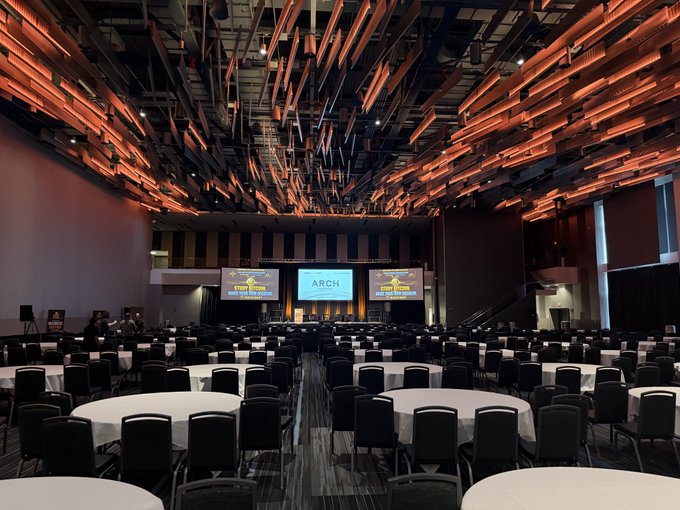 An empty conference hall or ballroom is set up with rows of round tables and chairs facing a stage with three projection screens, the center one displaying 'ARCH'.