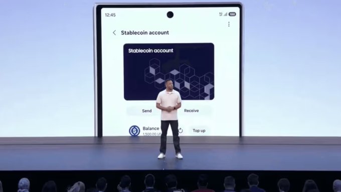 A man in a light pink shirt stands on a presentation stage in front of a large screen displaying a smartphone interface for a 'Stablecoin account', showing options to 'Send', 'Receive', and view a 'Balance' of '1,500.00 US'.