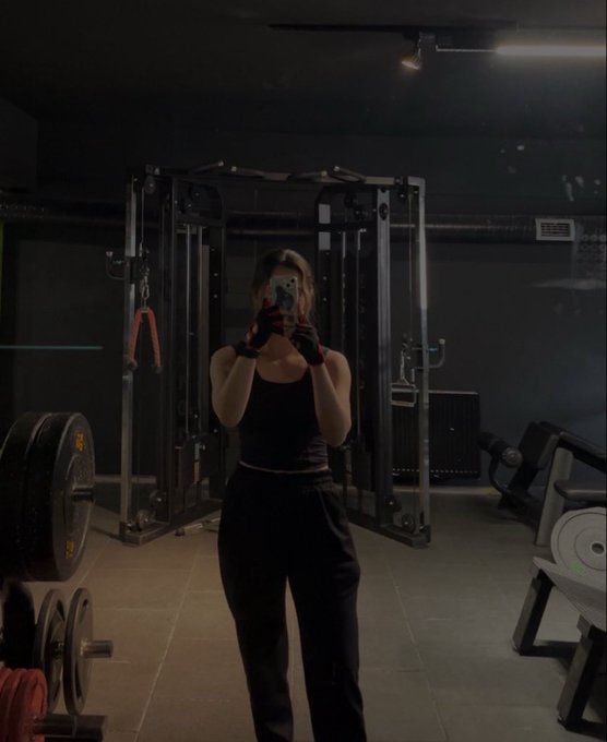 A woman in workout attire takes a mirror selfie in a dimly lit gym, with weight racks and exercise equipment visible around her.