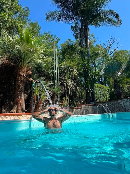 A man with tattoos washes his hair while standing in a bright turquoise swimming pool, surrounded by lush palm trees and tropical vegetation under a clear blue sky.
