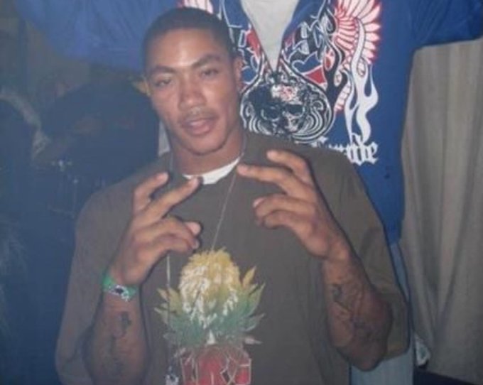A photo of a young man wearing a graphic t-shirt and chain necklace, making hand gestures, with other people visible in the background.
