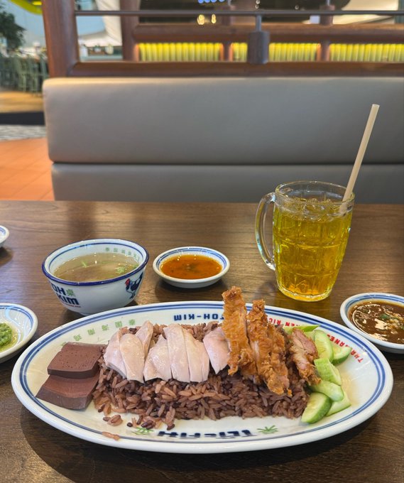 A plate of Hainanese chicken rice with cucumber slices, chicken, crispy skin, and pig's blood cubes sits alongside a bowl of clear broth, chili sauce, ginger paste, and dark soy sauce, with a glass mug of iced tea — the restaurant's name "WIH-HO" is visible on the plate rim, matching the "ct" (chicken rice) reference in the post.