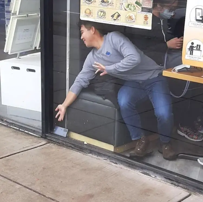 A man in a gray hoodie and jeans leans out of a store window, reaching down toward a smartphone lying on the sidewalk outside.