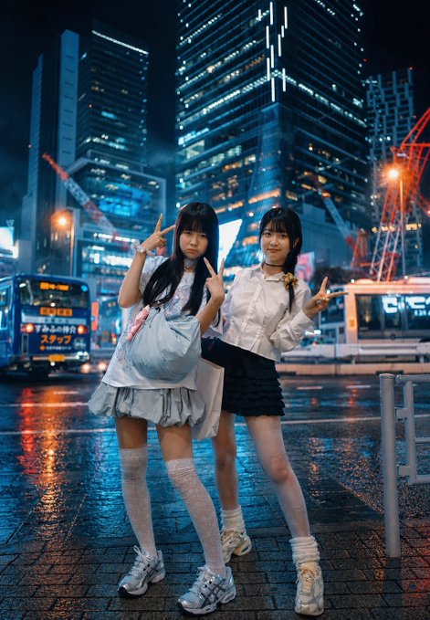 A nighttime portrait of two young women in coordinated white tops, skirts, and knee-high socks posing with peace signs in a rainy urban setting with illuminated skyscrapers and a bus in the background.