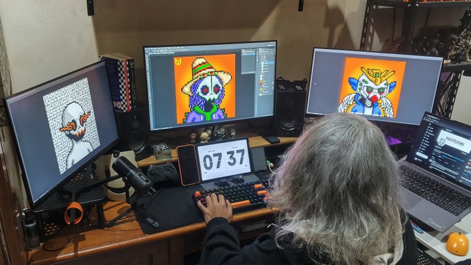 A person with long gray hair works at a multi-monitor computer setup, displaying various clown and character digital artworks, with a flip clock showing '07 37' visible on the desk.