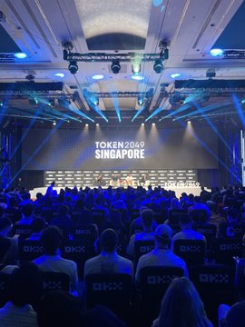 A photograph of the TOKEN2049 Singapore conference, showing a panel of speakers seated on stage in front of a large screen, with an audience seated in rows labeled 'OKX'.