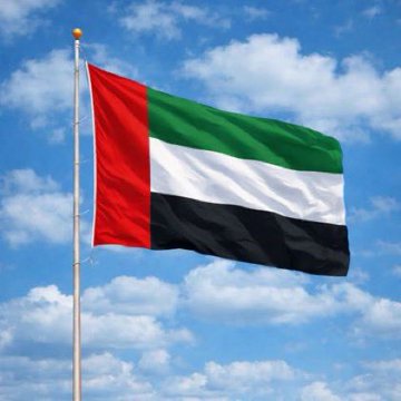 The flag of the United Arab Emirates waves on a flagpole against a blue sky with white clouds, featuring its characteristic vertical red stripe on the left and horizontal green, white, and black stripes.