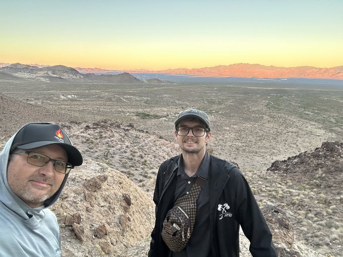 Two men wearing caps and glasses take a selfie in a desert landscape with mountains in the background, likely during sunrise or sunset given the warm lighting.