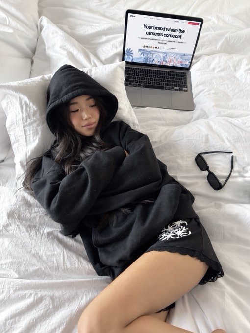 A young woman wearing a black hoodie lies on a white bed next to a laptop displaying an article titled 'Your brand where the cameras come out,' with sunglasses and earphones placed nearby.