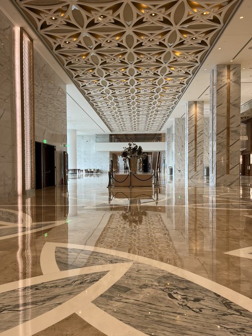 A grand, luxurious hotel lobby with an ornate geometric ceiling, polished marble floors, and elegant seating arranged around a floral centerpiece—likely the conference venue referenced in the post. The opulent interior contrasts with the post's mention of rough surrounding infrastructure and curiosity about attendance at the event.