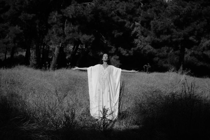 A black and white photograph shows a woman in a flowing white dress standing with her arms outstretched in a grassy field surrounded by trees.
