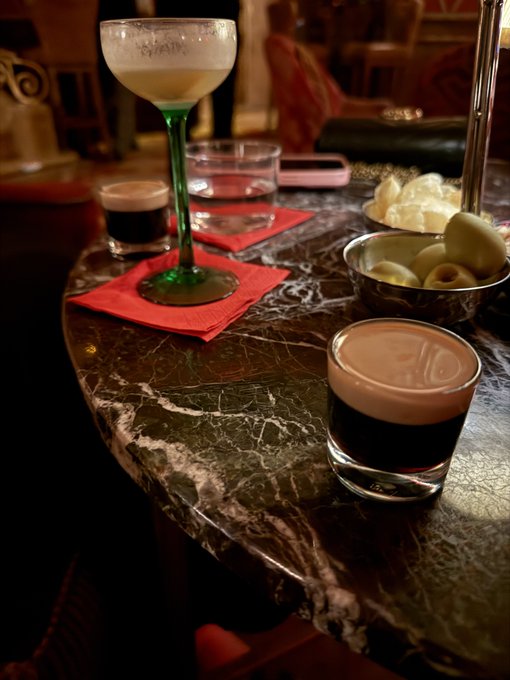 A dimly lit bar scene featuring a marble table topped with various cocktails, including one with a green stem, a small espresso-like drink, a glass of water, and bowls of olives and chips.