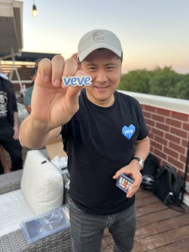 A smiling man wearing a VeVe-branded black t-shirt and gray cap proudly displays a "veve" sticker toward the camera at what appears to be an outdoor rooftop gathering, with a VeVe collectible case visible on the table behind him, capturing the community spirit of the Chicago VeVe meetup mentioned in the post.