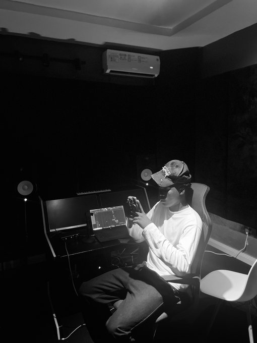 A black-and-white photo shows a young artist sitting in a dimly lit home studio wearing headphones, working at a laptop with a DAW (digital audio workstation) open on the screen, appearing engaged in music production. The image conveys the creative, behind-the-scenes process of beatmaking, aligning with the post's offer of a 20/80 split for label collaborations.